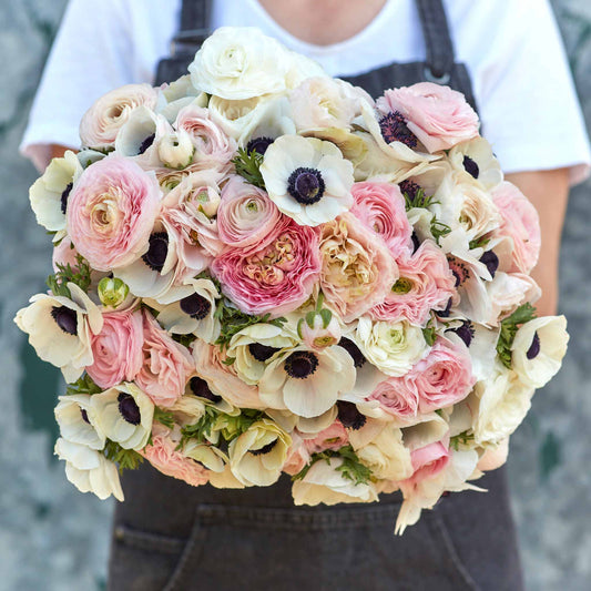A person holding a bouquet of Paris in Spring anemone and ranunculus flowers with a mix of pink, white, and beige petals.